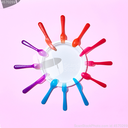 Image of Multicolored plastic utensil around white plate on pink.