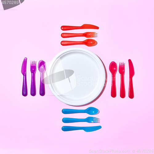Image of Multicolored plastic utensil around white plate on a light pink.