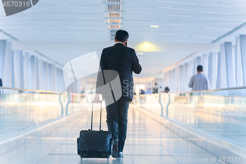 Image of Businessman walking and wheeling a trolley suitcase at the lobby, talking on a mobile phone. Business travel concept.