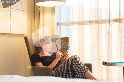 Image of Businesswoman on business travel, tired from business meeting, sitting alone in hotel room afterwork, reading and responding to emails and messages on her smart phone