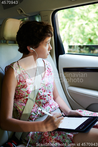 Image of Creative girl in earphones looking at car window and drawing on tablet during road trip