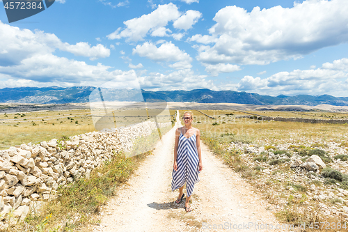 Image of Caucasian young woman in summer dress holding bouquet of lavender flowers while walking outdoor through dry rocky Mediterranean Croatian coast lanscape on Pag island in summertime