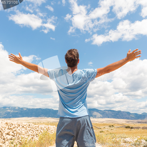 Image of Rear view of casual sporty man standing on a dirt country road rising hands up to the clouds on a blue summer sky. Freedom and travel adventure concept.