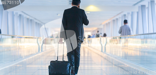 Image of Businessman walking and wheeling a trolley suitcase at the lobby, talking on a mobile phone. Business travel concept.