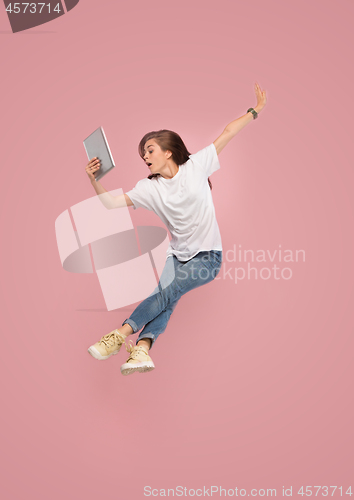 Image of Image of young woman over pink background using laptop computer or tablet gadget while jumping.