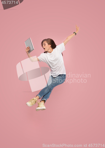 Image of Image of young woman over pink background using laptop computer or tablet gadget while jumping.