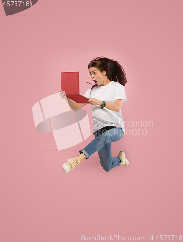 Image of Image of young woman over pink background using laptop computer or tablet gadget while jumping.