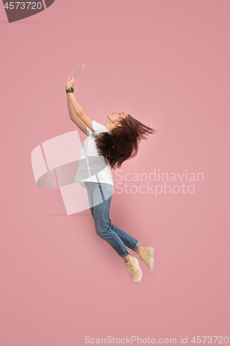 Image of Image of young woman over pink background using laptop computer or tablet gadget while jumping.