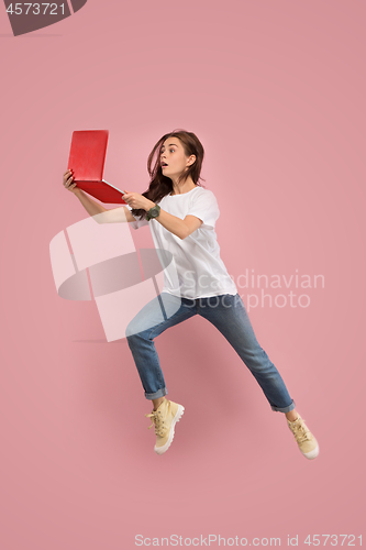 Image of Image of young woman over pink background using laptop computer or tablet gadget while jumping.