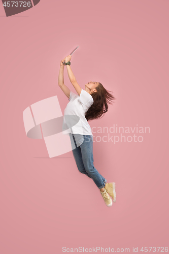 Image of Image of young woman over pink background using laptop computer or tablet gadget while jumping.