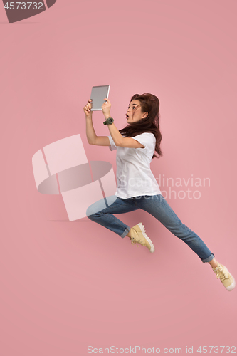 Image of Image of young woman over pink background using laptop computer or tablet gadget while jumping.