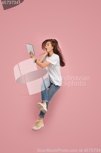 Image of Image of young woman over pink background using laptop computer or tablet gadget while jumping.