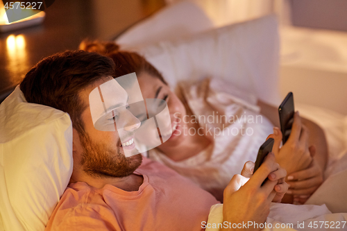 Image of happy couple using smartphones in bed at night