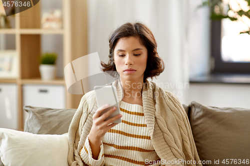 Image of sad woman in blanket using smartphone at home