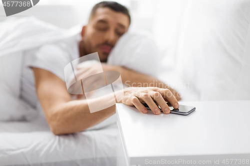 Image of sleepy young man reaching for smartphone in bed