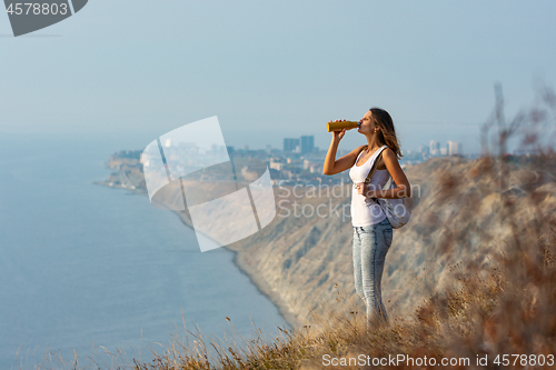 Image of Girl drinks from a bottle of water, standing against the backdrop of a mountain and sea landscape