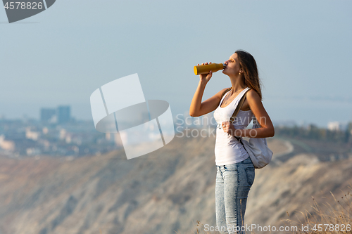 Image of Girl drinks from a bottle of water, standing against the backdrop of a mountain and city landscape