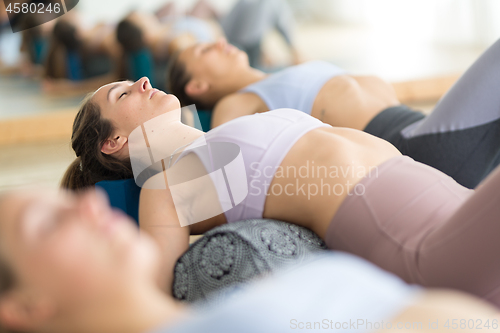 Image of Restorative yoga with a bolster. Group of three young sporty attractive women in yoga studio, lying on bolster cushion, stretching and relaxing during restorative yoga. Healthy active lifestyle