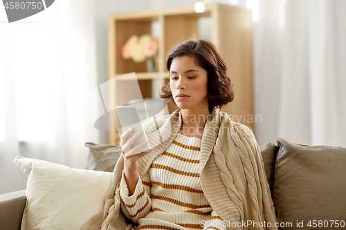 Image of sad woman in blanket using smartphone at home