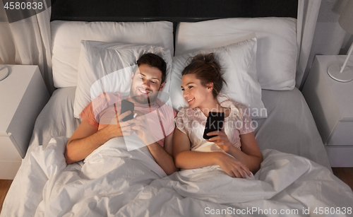 Image of happy couple using smartphones in bed at night