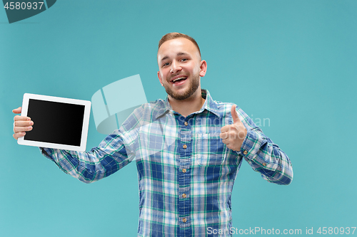 Image of Studio picture of positive man isolated on blue background standing in casual clothes holding tablet and showing it blank screen with happy smile