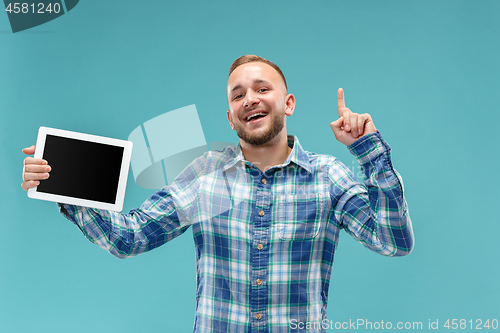 Image of Studio picture of positive man isolated on blue background standing in casual clothes holding tablet and showing it blank screen with happy smile