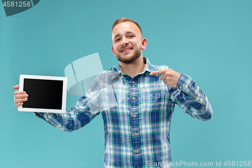 Image of Studio picture of positive man isolated on blue background standing in casual clothes holding tablet and showing it blank screen with happy smile