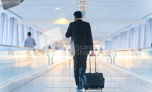 Image of Businessman walking and wheeling a trolley suitcase at the lobby, talking on a mobile phone. Business travel concept.