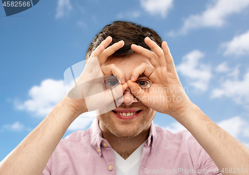 Image of young man looking through finger glasses over sky