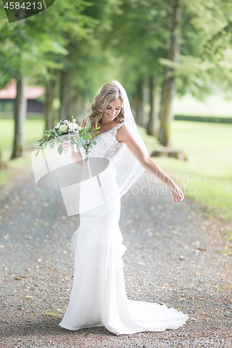 Image of Full length portrait of beautiful sensual young blond bride in long white wedding dress and veil, holding bouquet outdoors in natural background