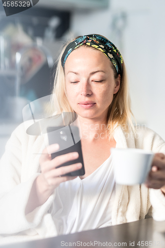 Image of Beautiful caucasian woman at home, feeling comfortable wearing white bathrobe, taking some time to herself, drinking morning coffee and reading news on mobile phone device in the morning