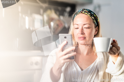 Image of Beautiful caucasian woman at home, feeling comfortable wearing white bathrobe, taking some time to herself, drinking morning coffee and reading news on mobile phone device in the morning