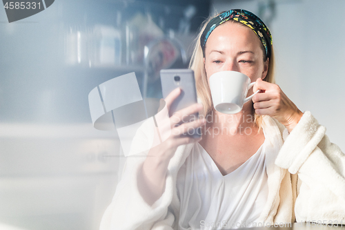 Image of Beautiful caucasian woman at home, feeling comfortable wearing white bathrobe, taking some time to herself, drinking morning coffee and reading news on mobile phone device in the morning