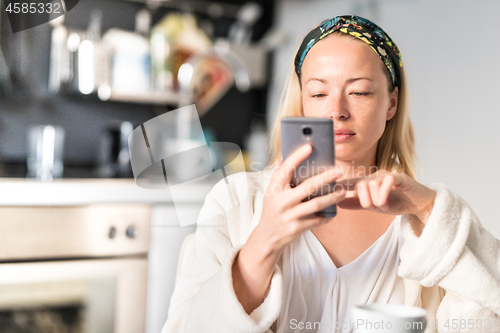 Image of Beautiful caucasian woman at home, feeling comfortable wearing white bathrobe, taking some time to herself, drinking morning coffee and reading news on mobile phone device in the morning