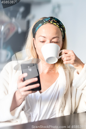 Image of Beautiful caucasian woman at home, feeling comfortable wearing white bathrobe, taking some time to herself, drinking morning coffee and reading news on mobile phone device in the morning