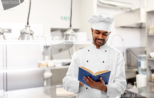 Image of happy indian chef reading cookbook at kitchen