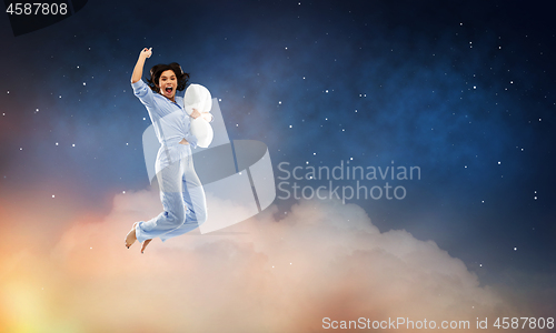 Image of happy woman in blue pajama jumping with pillow