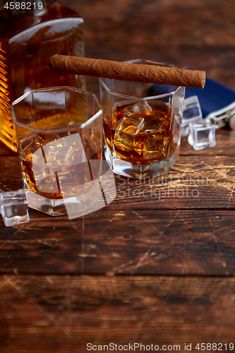 Image of Bottle of whiskey with two glasses and cuban cigar placed on rustic wooden table