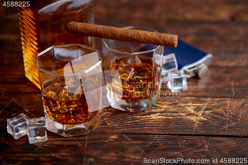 Image of Bottle of whiskey with two glasses and cuban cigar placed on rustic wooden table