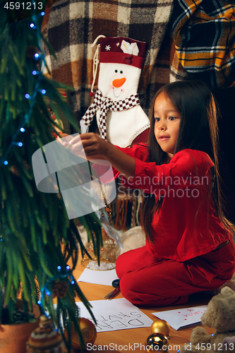 Image of christmas, holidays and childhood concept - happy girl in red clothes decorating natural fir tree
