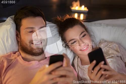 Image of happy couple using smartphones in bed at night