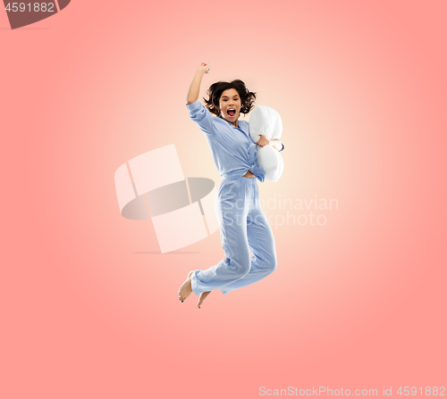 Image of happy woman in blue pajama jumping with pillow