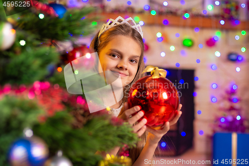 Image of Happy girl peeps out from behind a beautiful Christmas tree with a ball in her hands