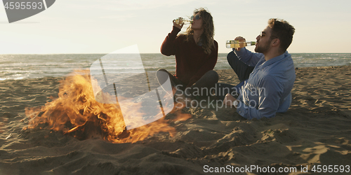 Image of Loving Young Couple Sitting On The Beach beside Campfire drinkin