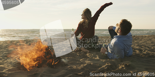 Image of Loving Young Couple Sitting On The Beach beside Campfire drinkin