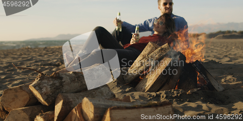 Image of Loving Young Couple Sitting On The Beach beside Campfire drinkin