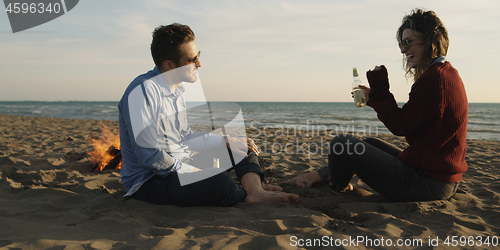 Image of Loving Young Couple Sitting On The Beach beside Campfire drinkin