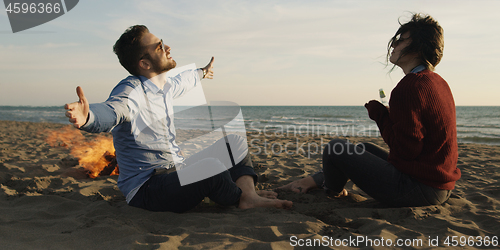 Image of Loving Young Couple Sitting On The Beach beside Campfire drinkin