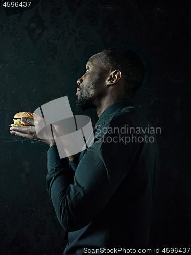 Image of young african american man eating hamburger and looking away on black studio