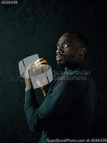 Image of young african american man eating hamburger and looking away on black studio